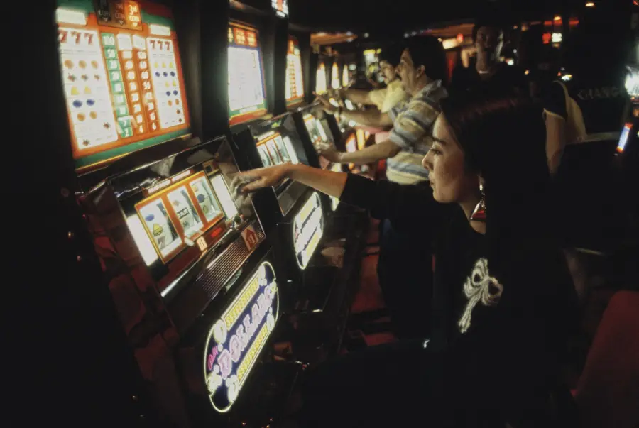 A woman sits at a row of slot machines in a dimly lit casino, reaching to pull the lever. Other people are playing machines nearby. The atmosphere appears lively and focused.