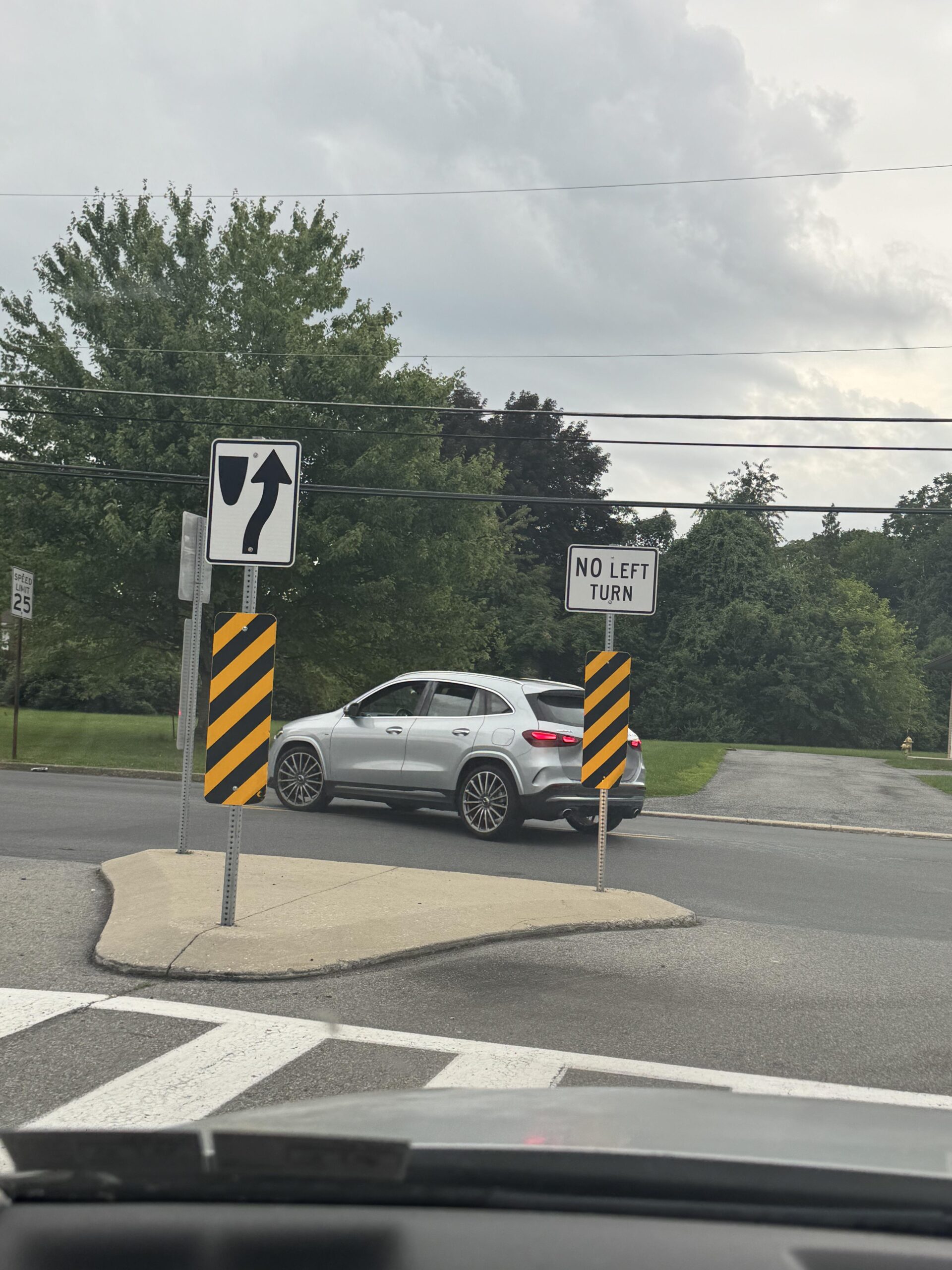 A silver car turns left at an intersection despite a posted sign that says “No Left Turn.” The weather is cloudy and trees are in the background.