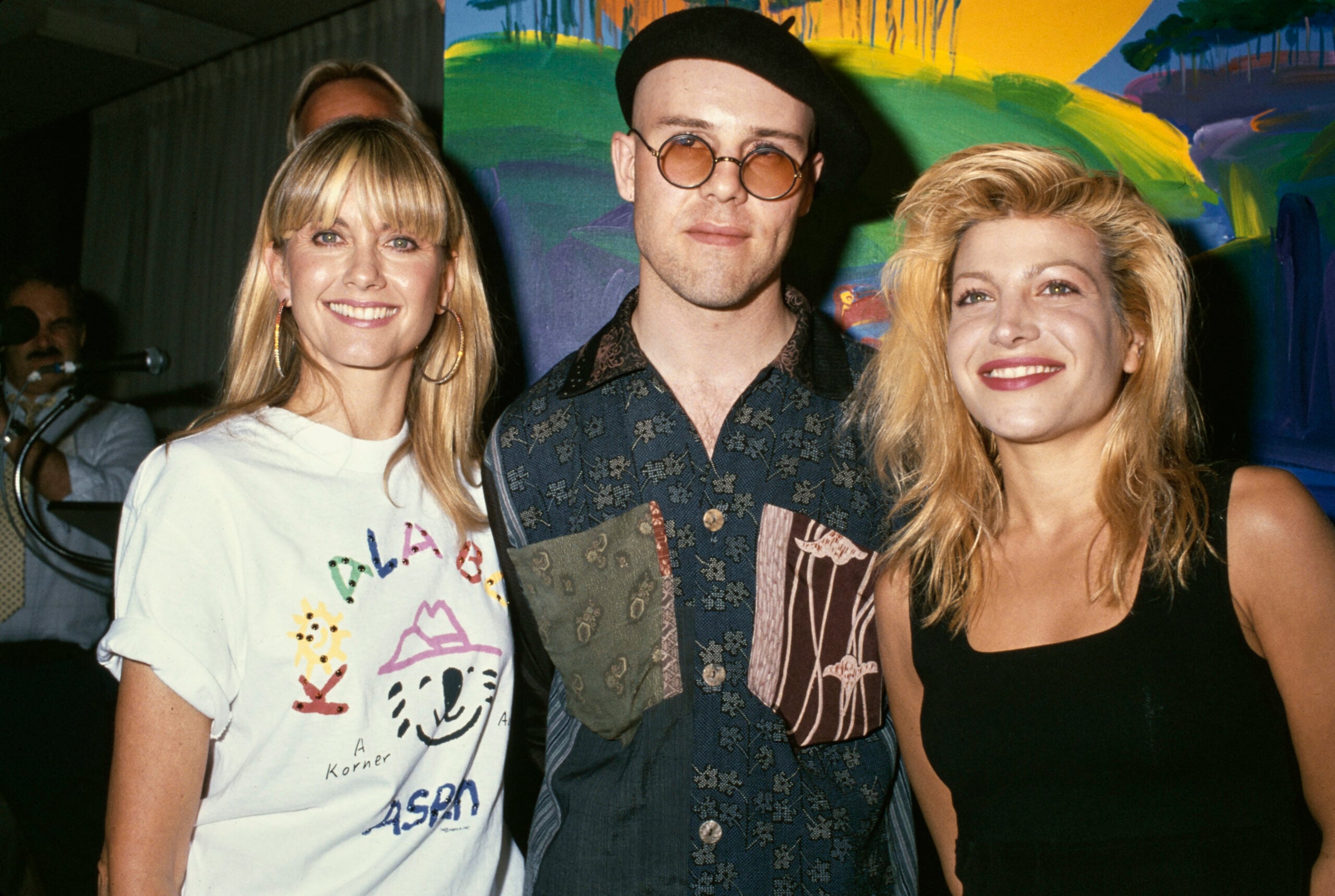 Three people pose together, smiling, in front of a colorful painted background. The person in the middle wears round glasses and a hat, while the others have long blonde hair and casual clothing.