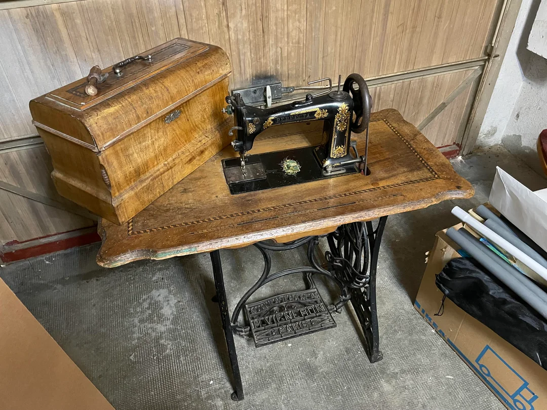 An antique black sewing machine with gold details sits on a wooden table. A wooden case is placed next to it. The setup is in a garage or workshop, with boxes and pipes visible nearby.