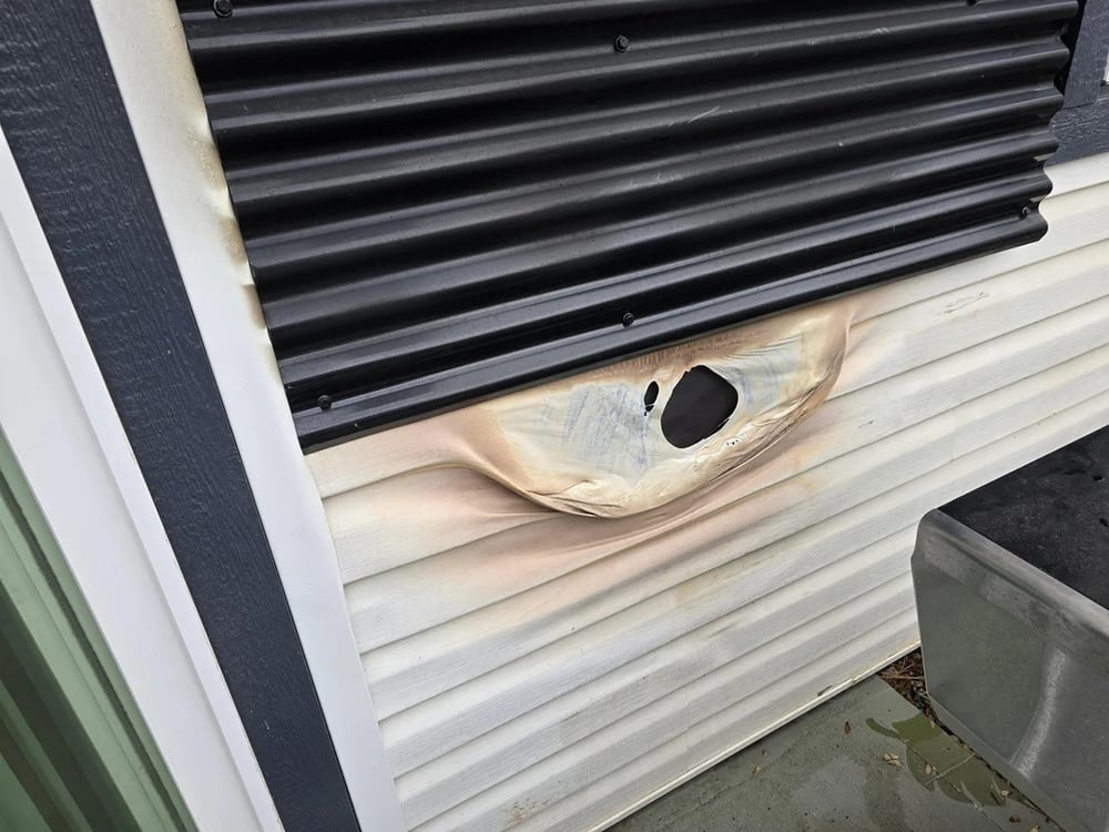 A section of white plastic siding on a building is melted and warped beneath a black metal window shutter, with a large hole in the center of the damage.