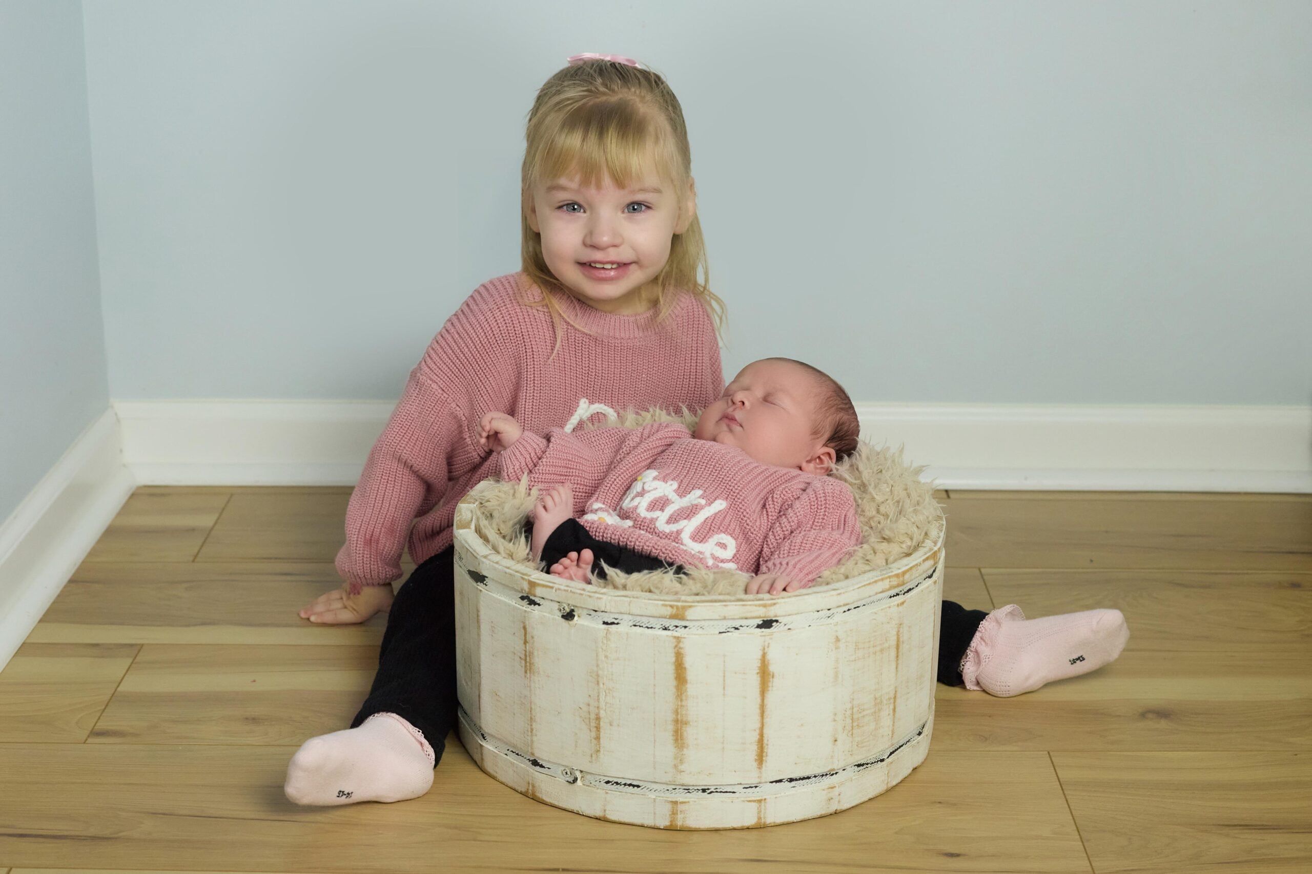 A young child sits on a wooden floor, smiling, beside a newborn baby lying in a round, white wooden basket. Both wear pink sweaters and black pants. The background is a light blue wall.