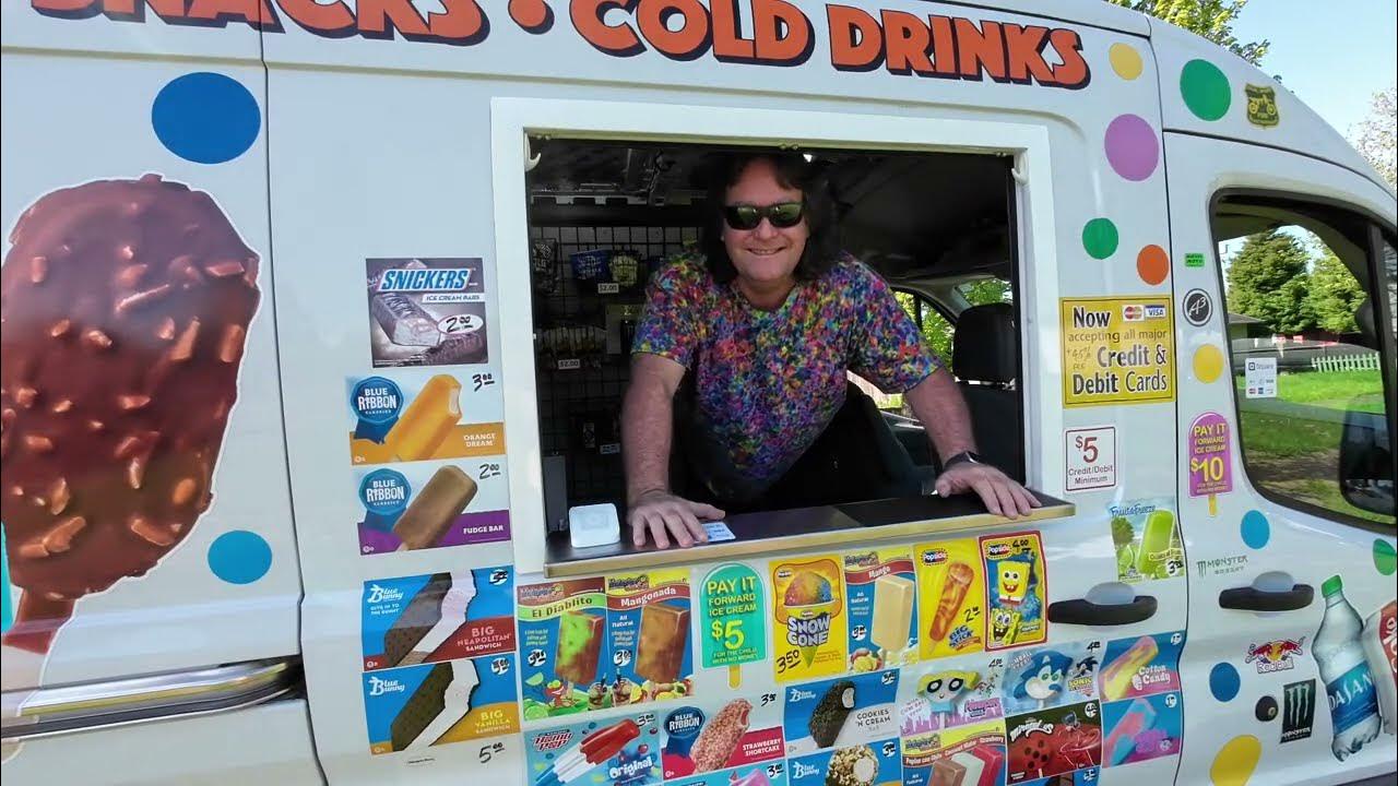 A person wearing sunglasses and a colorful shirt leans out of an ice cream truck window, surrounded by images of ice creams, snacks, and price signs on the truck.
