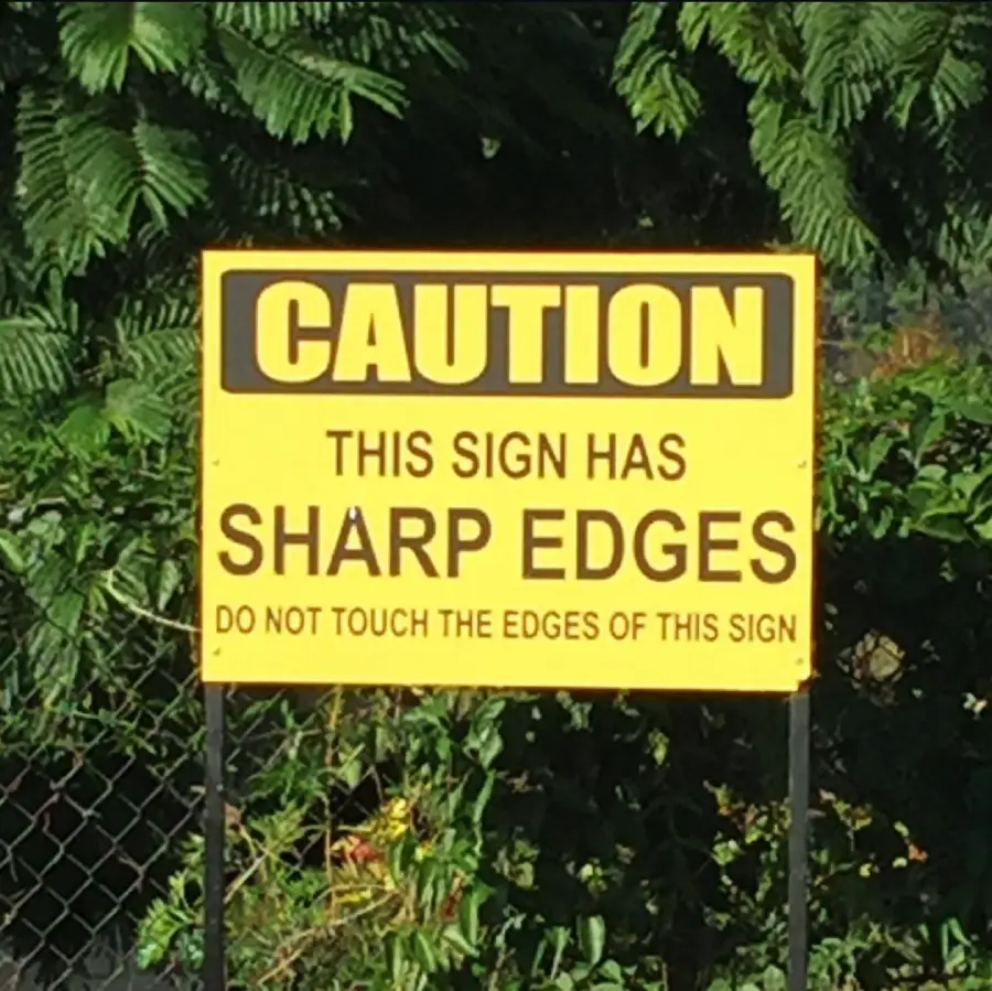 Yellow caution sign reads: “CAUTION. THIS SIGN HAS SHARP EDGES. DO NOT TOUCH THE EDGES OF THIS SIGN.” Green foliage is visible in the background.