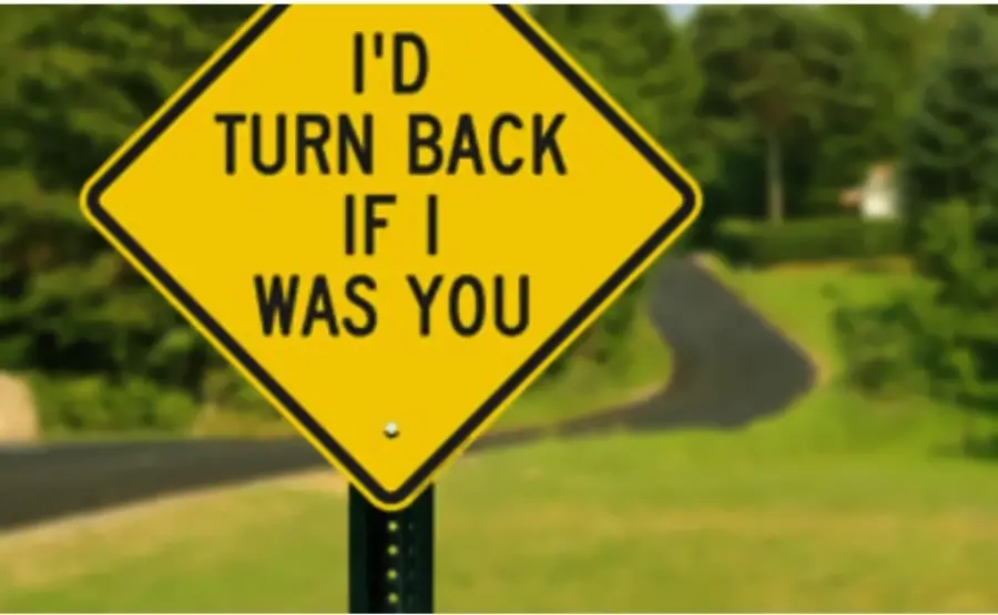 Yellow road sign reads, “I’D TURN BACK IF I WAS YOU,” with a blurred rural road and green trees in the background.