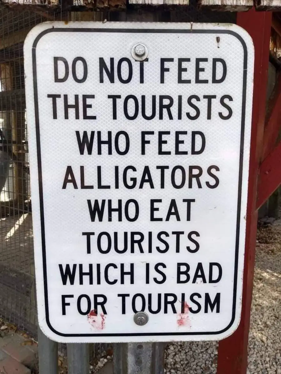 A sign reads: “DO NOT FEED THE TOURISTS WHO FEED ALLIGATORS WHO EAT TOURISTS WHICH IS BAD FOR TOURISM.” The sign is posted outdoors near a fence and gravel.