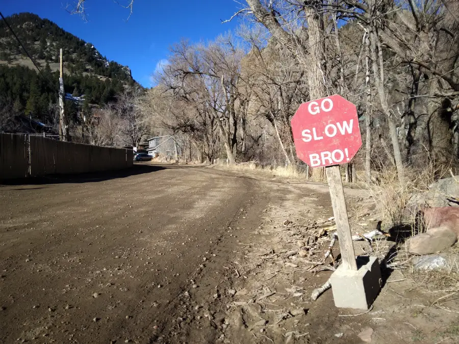 A red, octagonal sign reads “GO SLOW BRO!” beside a dirt road lined with leafless trees and mountains in the background under a clear blue sky.