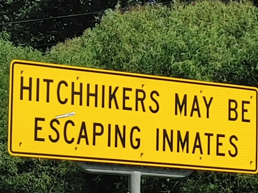 A yellow road sign reads, “HITCHHIKERS MAY BE ESCAPING INMATES,” in black capital letters. Trees and power lines are visible in the background.