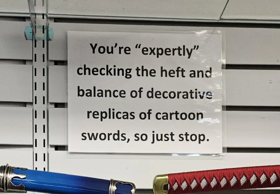 A sign reads: “You’re ‘expertly’ checking the heft and balance of decorative replicas of cartoon swords, so just stop.” Decorative swords are visible below the sign.