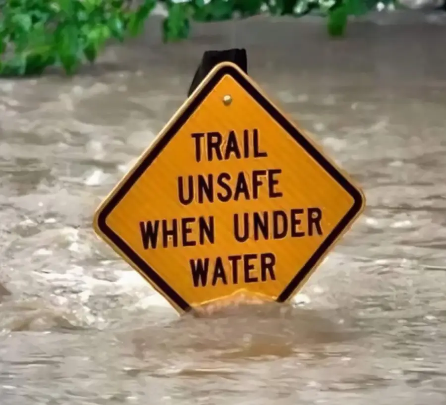 A yellow sign reading “Trail unsafe when under water” is partially submerged in floodwaters, with tree branches visible above.