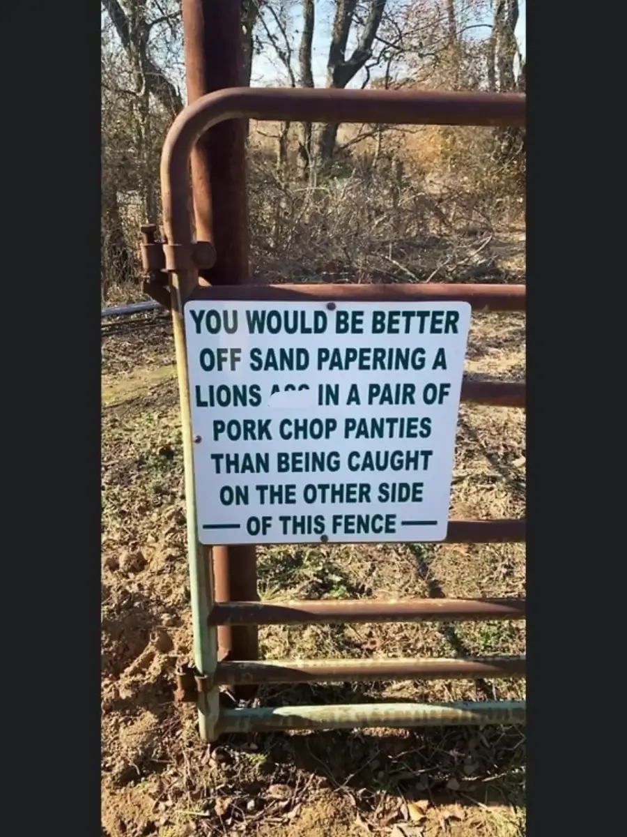 A metal fence gate with a humorous warning sign that reads, “You would be better off sandpapering a lion’s ass in a pair of pork chop panties than being caught on the other side of this fence.”