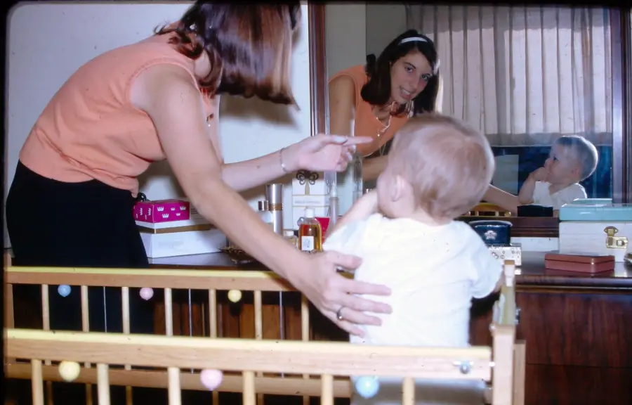 A woman leans over a wooden crib, gesturing to a toddler who faces away. Their reflection is visible in a mirror behind them.