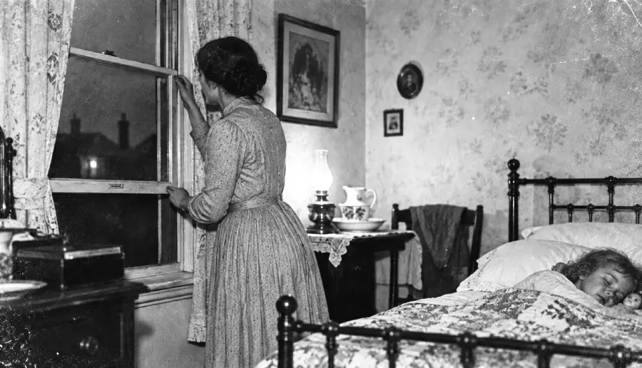 A woman in old-fashioned clothing closes a window at night while a child sleeps in bed. The room is softly lit by a lamp and decorated with floral wallpaper, framed pictures, and vintage furniture.