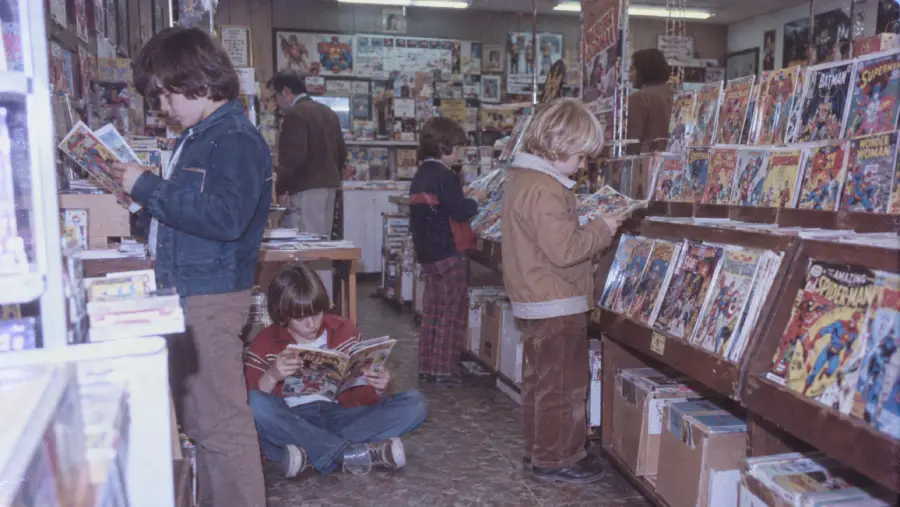 Four children read comic books in a store aisle lined with colorful comics, while adults browse in the background. The floor is tiled and the store appears busy and full of graphic novels and posters.