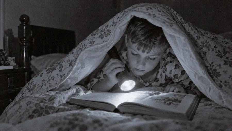 A child reads a book under a blanket at night, using a flashlight for light. The scene is cozy, with the child fully focused on the book and surrounded by patterned bedding.