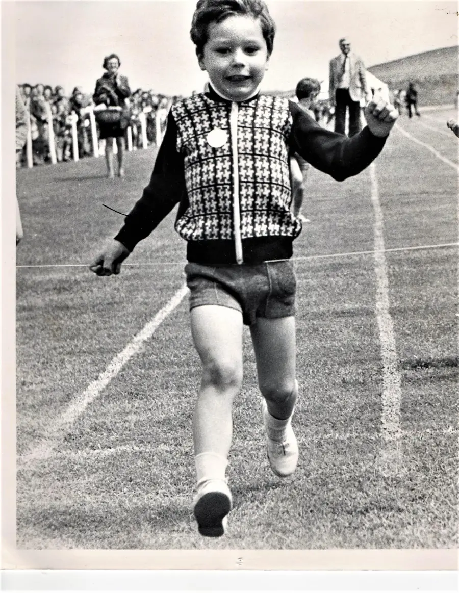 A young child crosses a finish line on a running track, smiling and wearing a patterned vest and shorts. Spectators stand in the background on a grassy field.