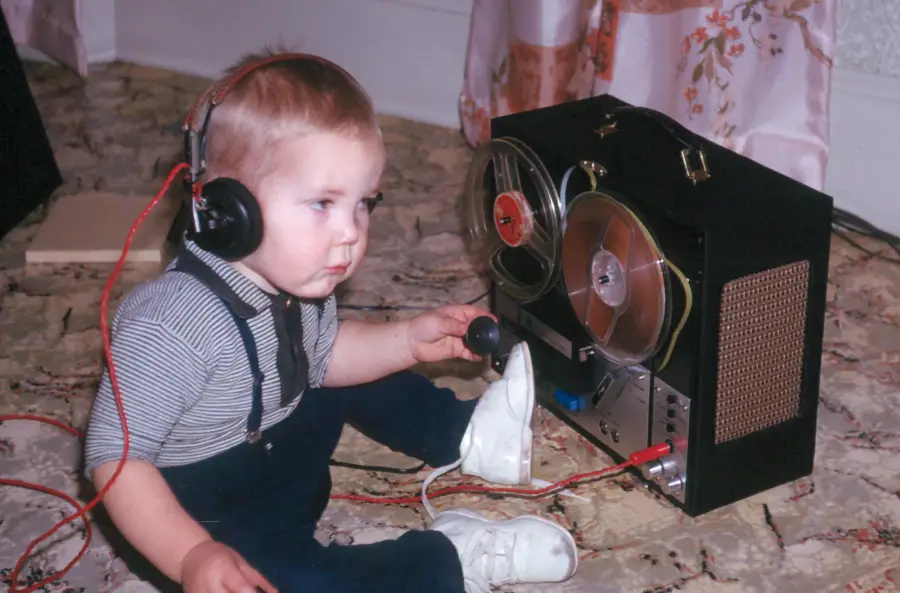 A young child wearing headphones sits on a carpeted floor and listens to a reel-to-reel tape recorder, holding a microphone and appearing focused.