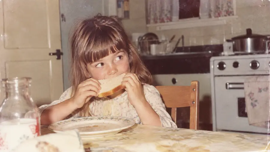 A young girl with brown hair and bangs sits at a kitchen table, eating a sandwich. An empty plate is in front of her. The kitchen has a stove, dishes, and a floral curtain on the window in the background.