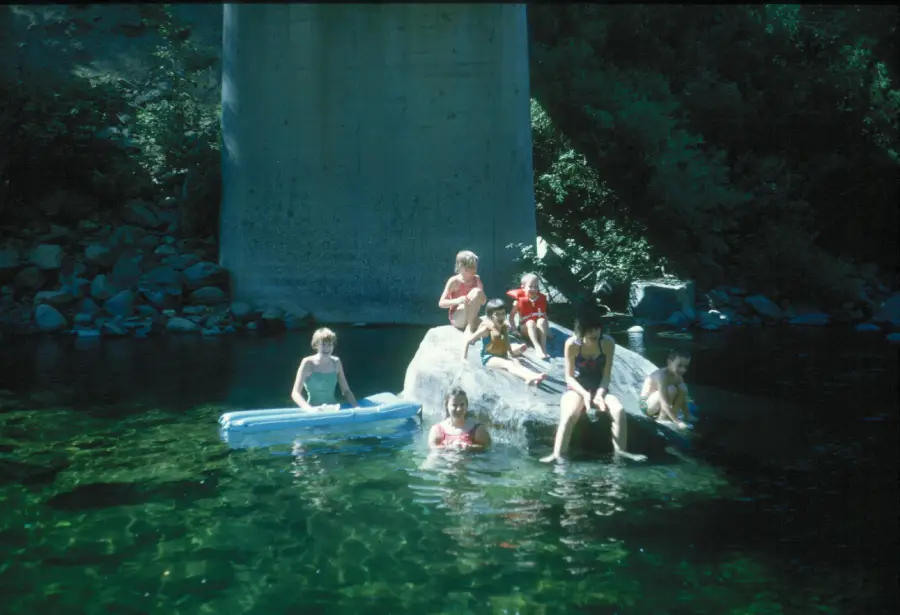 Six children play in a clear river under a bridge. Some sit on a large rock, while others are in the water or on a blue float. Sunlight filters through the trees, illuminating the scene.