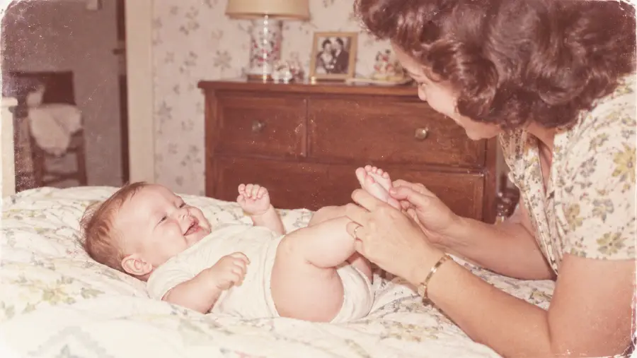 A woman smiles and plays with a laughing baby lying on a bed in a cozy, softly lit bedroom. A wooden dresser with family photos and a lamp is visible in the background.