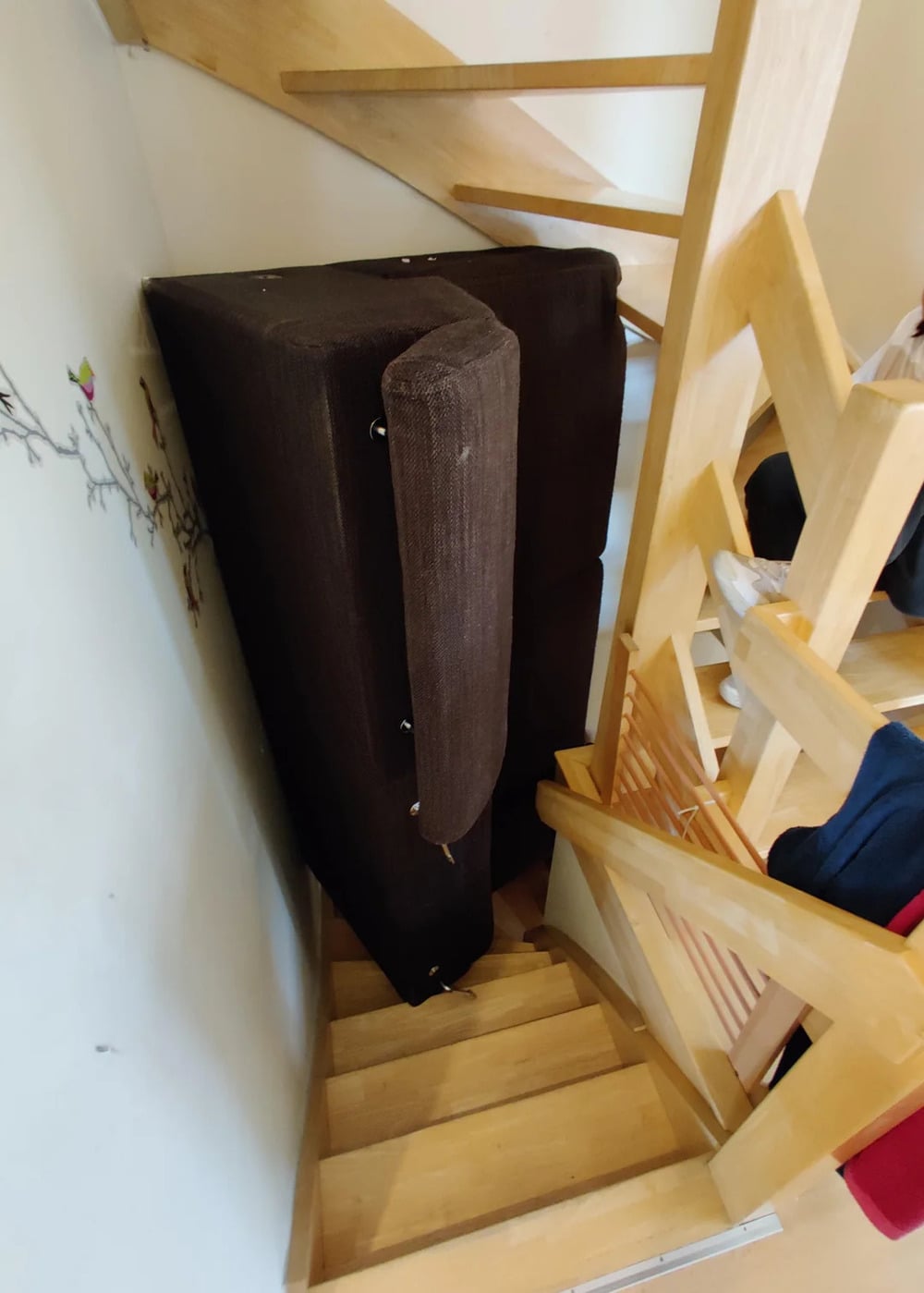 A brown couch is stuck upright in a tight corner on a wooden staircase, wedged between the stair railings and the wall, making it difficult to move either up or down the stairs.