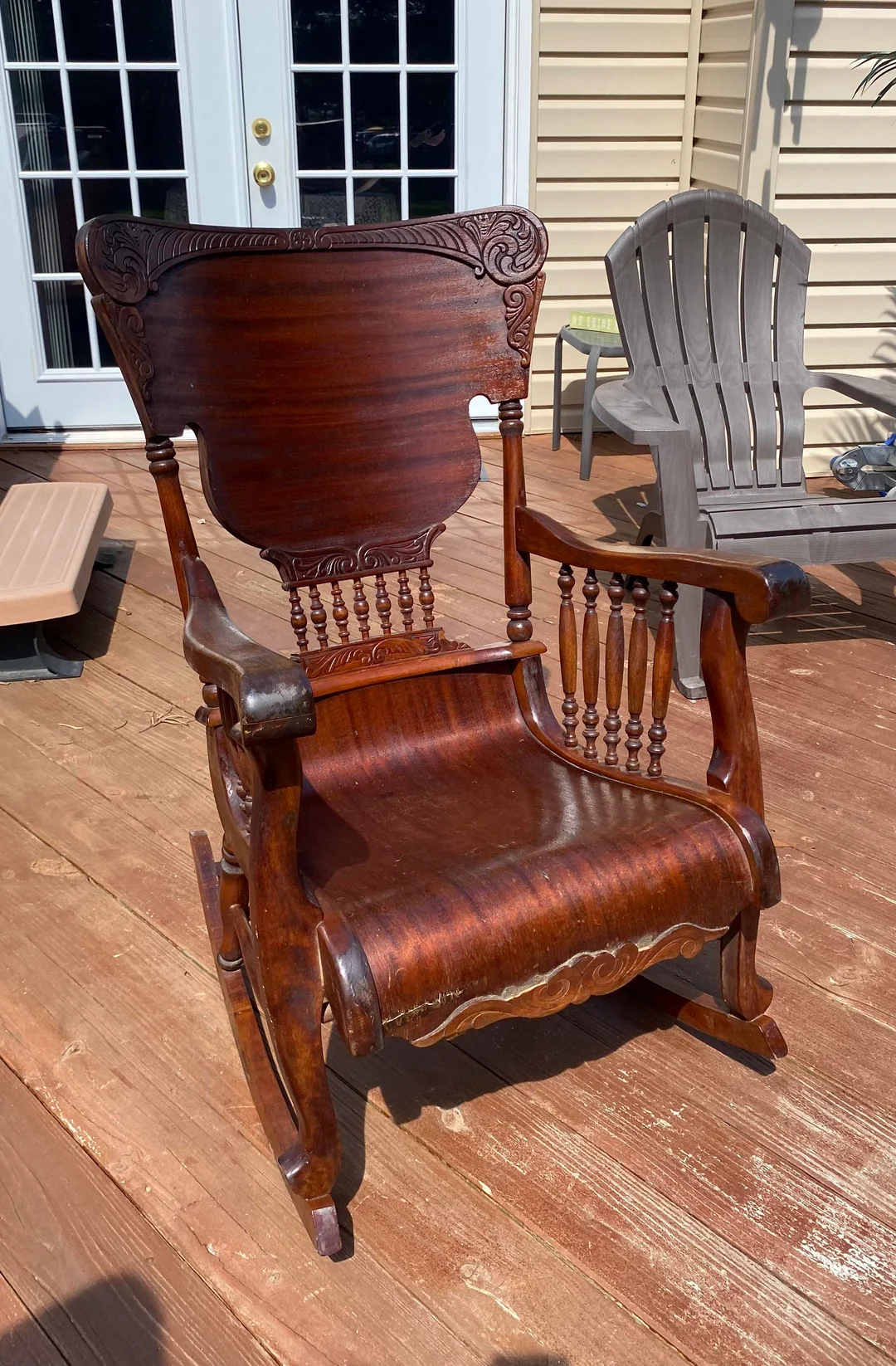 Antique wooden rocking chair with ornate carvings sits on a sunlit wooden deck, with patio furniture and sliding glass doors visible in the background.
