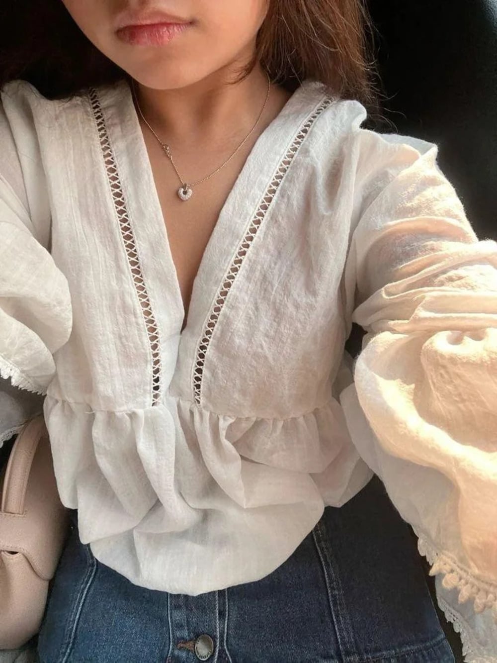 A person wearing a white, long-sleeved, v-neck blouse with lace trim and blue jeans, holding a light-colored bag. Their face is partially visible.