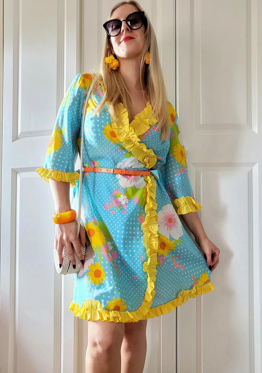 Woman in sunglasses wearing a blue dress with yellow and white floral print, yellow ruffle trim, orange earrings, bracelet, and carrying a white purse, standing in front of white doors.