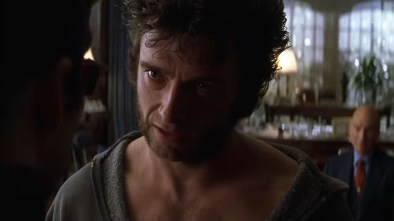 A man with messy hair and sideburns looks intensely at someone off-screen; another older man sits in the background, blurred, in a well-lit room.
