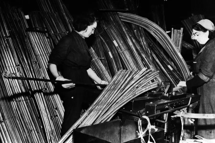 Two women work with bundles of long metal tubes in a factory setting. One woman arranges the tubes while the other operates a machine, with more stacked tubes in the background. The scene appears industrial and historical.