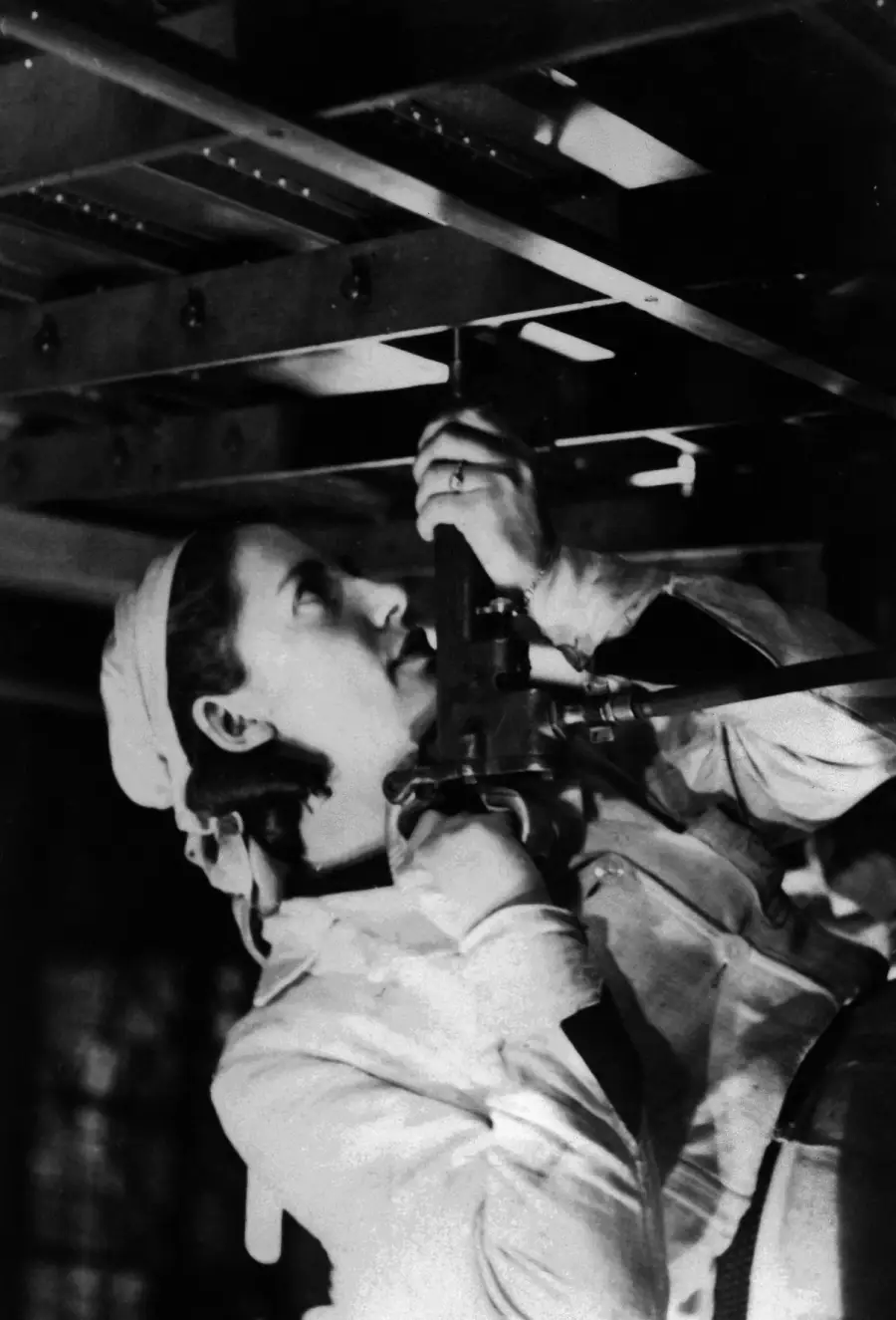 A woman wearing a headscarf and work gloves uses a tool to assemble part of a metal structure, looking focused while working beneath beams. The image is in black and white.