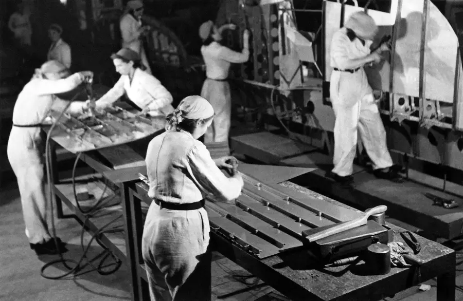 Women in uniforms work on assembling aircraft parts at a factory during World War II, using tools and machinery at workbenches and large structures in the industrial setting.