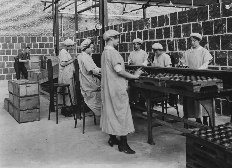 Six women in uniforms and caps work at a factory assembly line with jars. A man stands in the background near wooden crates. The setting appears industrial with brick and stone walls.