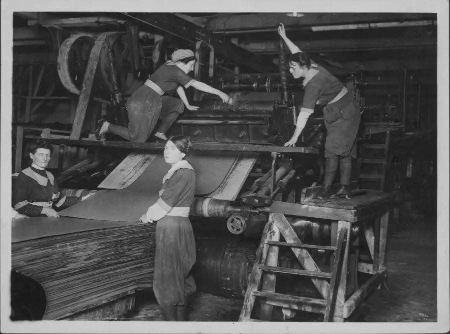 Four women operate a large industrial machine, handling sheets of material in a factory setting, with two women working atop the machinery and two standing beside or in front of it.