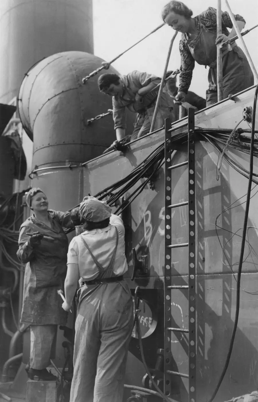 Four women in work overalls are repairing or assembling machinery on an industrial site, possibly a shipyard or factory, with pipes and cables visible in the background.