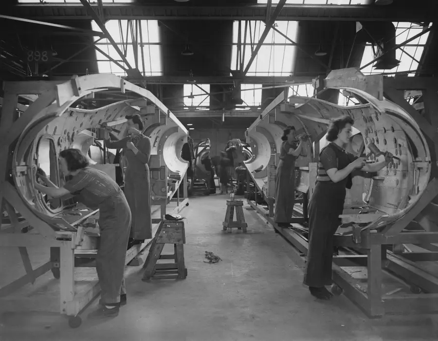 Women work on large cylindrical aircraft fuselages in a factory, wearing uniforms and using tools. The factory has high ceilings with windows letting in natural light.
