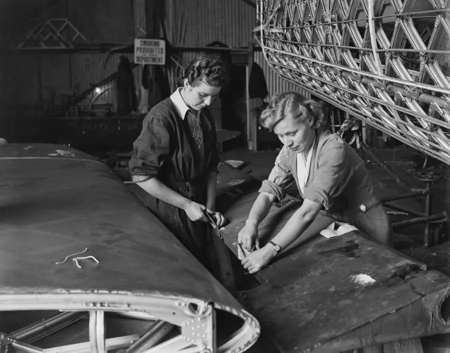 Two women work together assembling or repairing part of an aircraft wing in an industrial workshop, surrounded by metal framework and machinery.