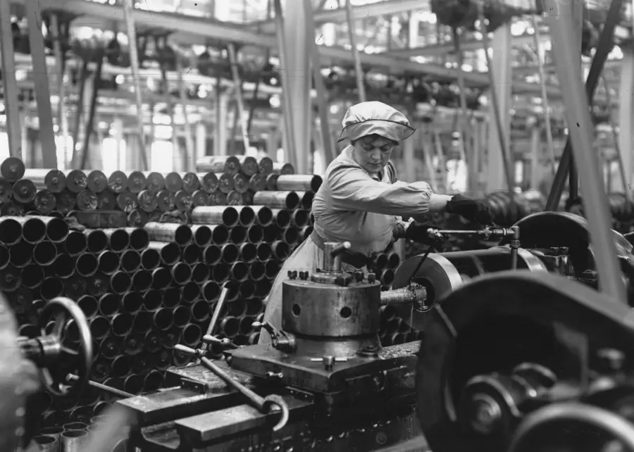 A woman in work clothes operates machinery in a factory filled with metal cylinders, likely during World War II, highlighting industrial labor and wartime production efforts.