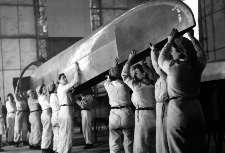 A group of women in uniform work together to lift a large section of an aircraft wing inside a hangar.