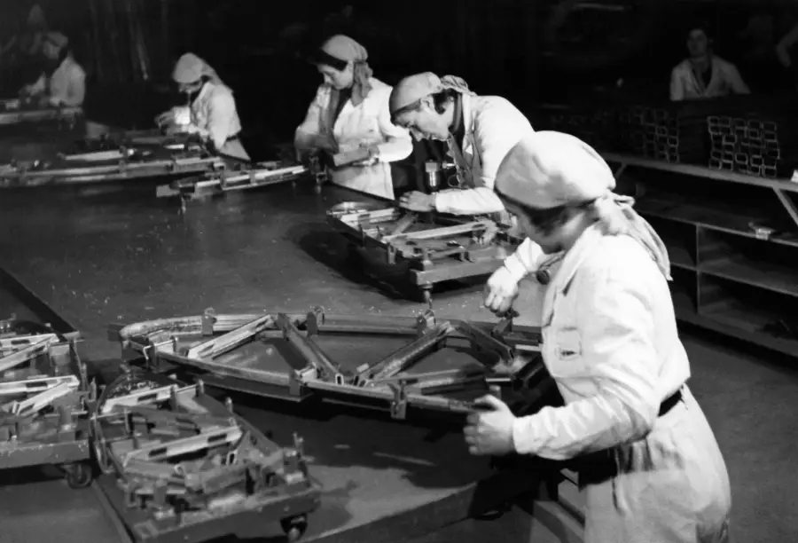 Women in uniforms and headscarves work at a factory assembly line handling metal frame parts, focusing intently on their tasks in a busy industrial setting.