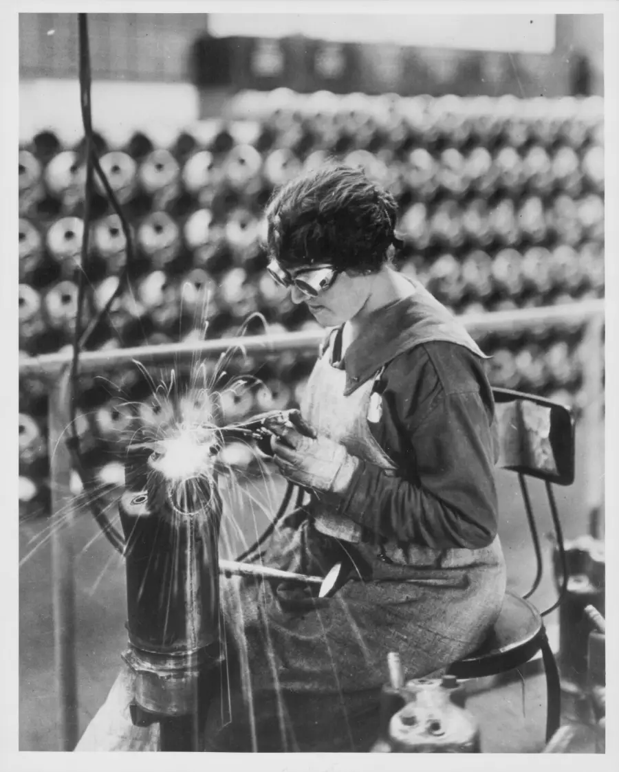 A woman wearing protective goggles welds metal while seated, surrounded by industrial equipment in a factory setting. Sparks fly from her work, and spools of material are visible in the background.