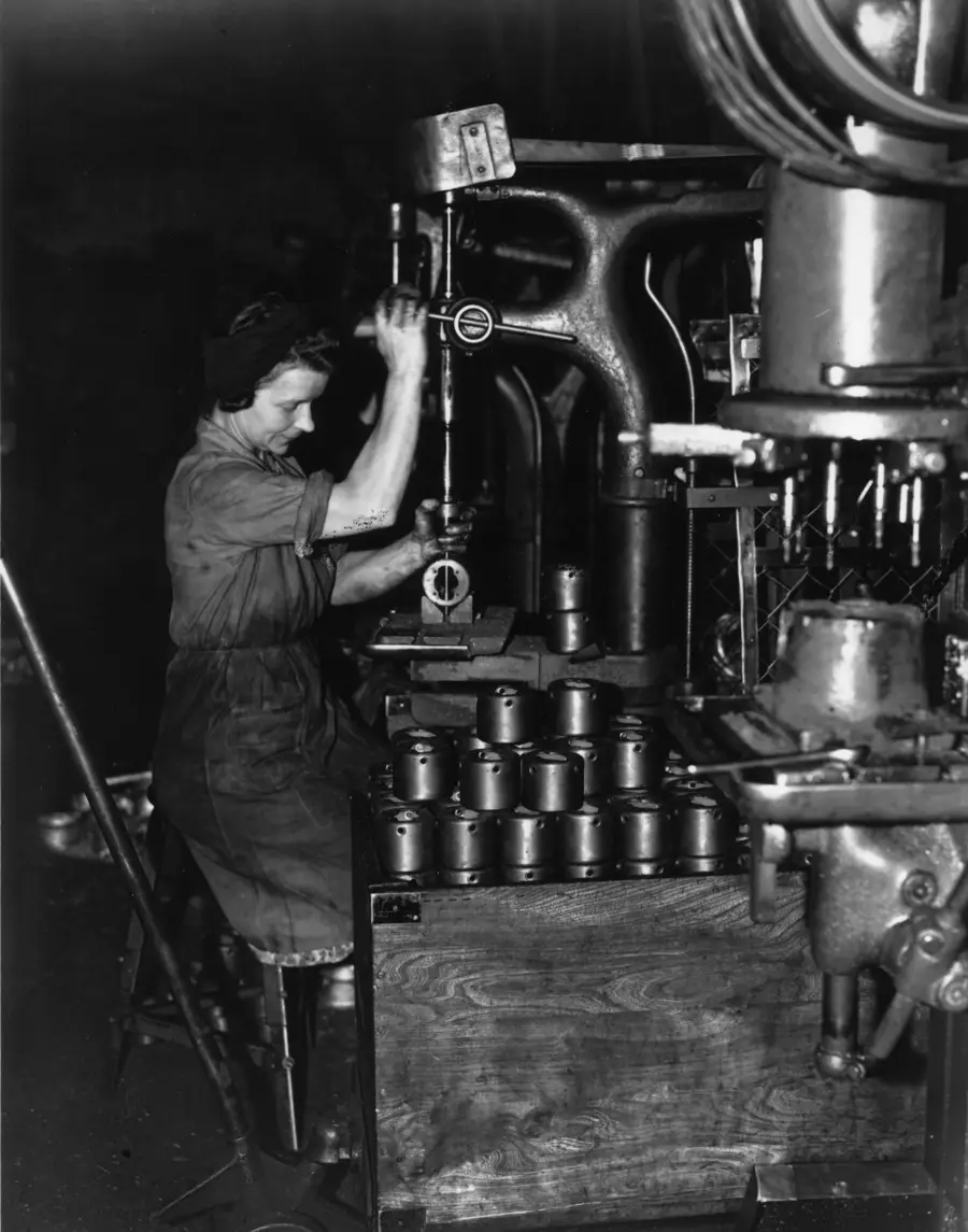 A woman in work clothes uses a drilling machine in a factory, surrounded by metal parts on a table. The scene is black and white, suggesting it is from a historical period, likely during wartime industrial production.