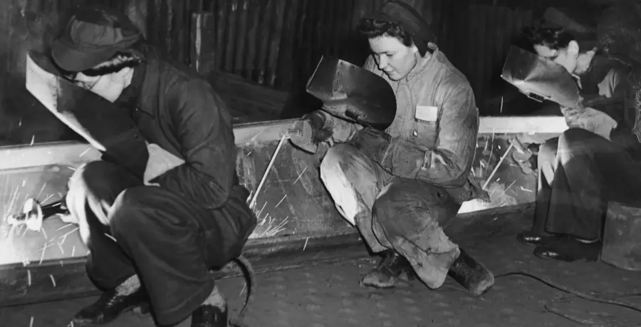 Three women in work clothes and protective masks kneel and weld metal beams, with sparks flying, in a black-and-white photo, likely from a mid-20th-century factory or industrial setting.