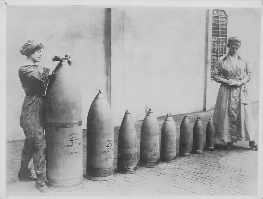 Two women in early 20th-century clothing stand by a row of large artillery shells arranged by size. One woman works on the largest shell, while the other writes on a clipboard.