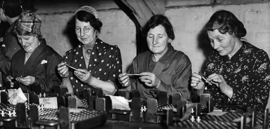 Four women sit in a row at a workbench assembling small parts, possibly in a factory setting. They are focused on their tasks, wearing work clothes, with trays and equipment in front of them.