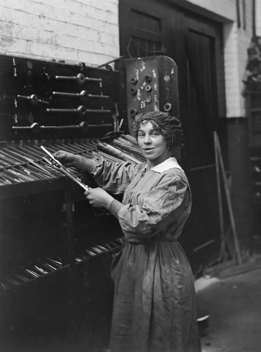 A woman in a long dress and cap stands in front of a tool rack, holding a metal rod in one hand and looking at the camera, in a workshop setting.
