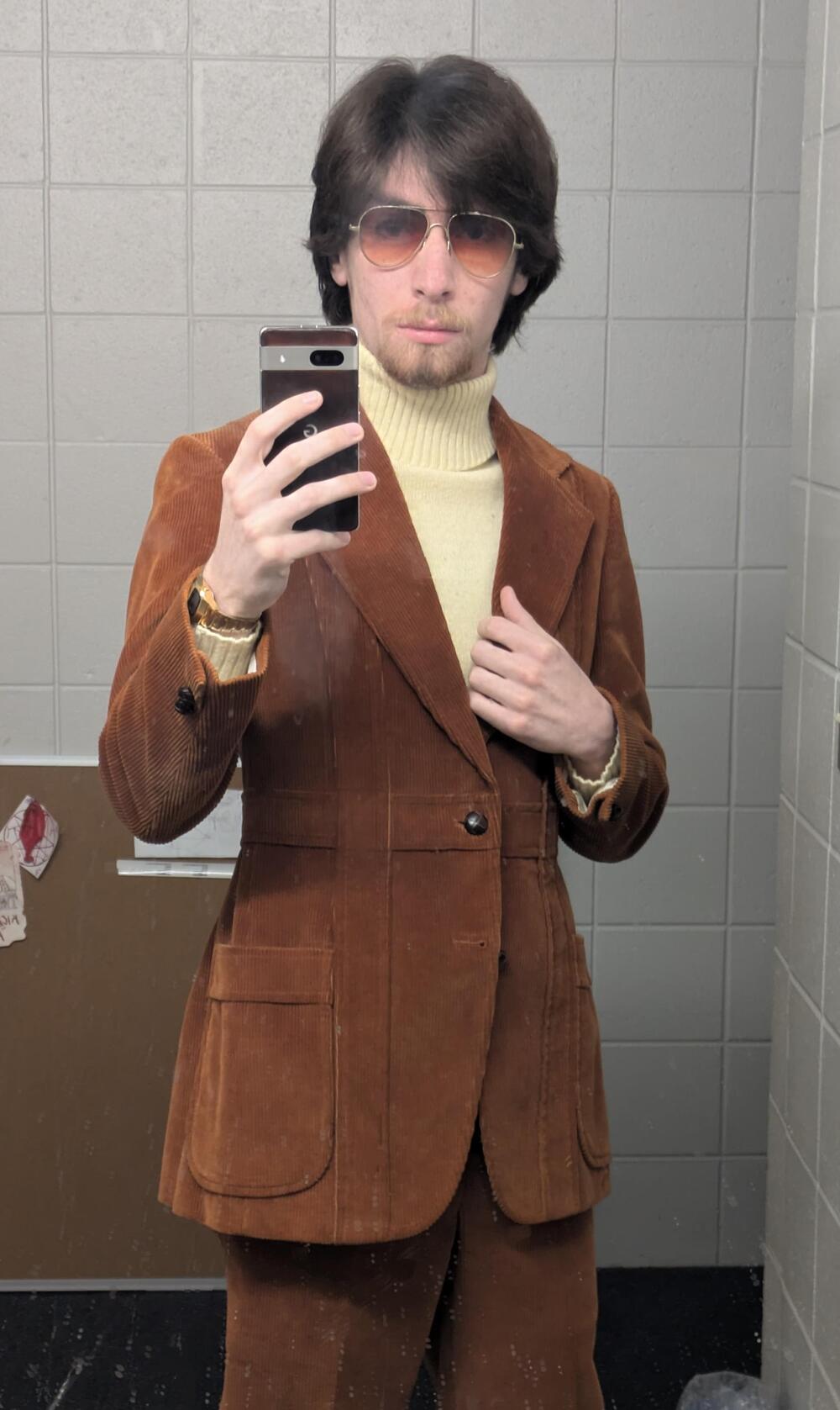 A person wearing a brown corduroy suit, cream turtleneck, and sunglasses takes a mirror selfie in a tiled bathroom.