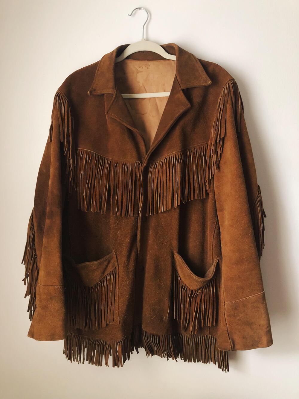 Brown suede jacket with fringe detailing on the chest, sleeves, and pockets, hanging on a white hanger against a plain white wall.