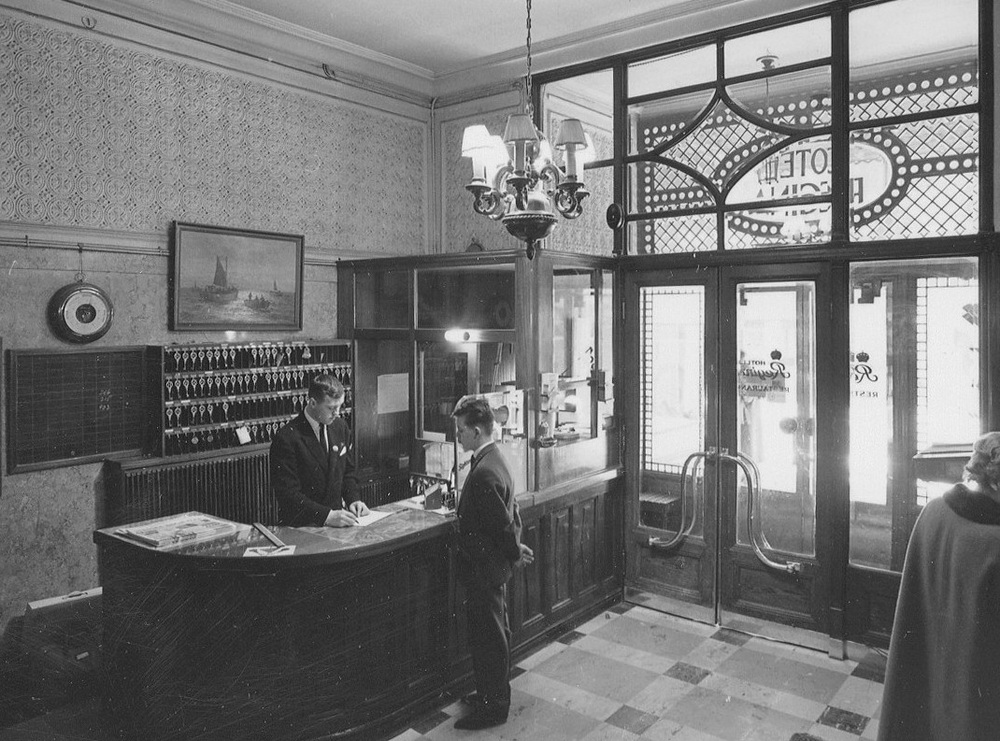 A hotel receptionist writes in a ledger at a wooden front desk while a guest stands opposite him, in a lobby with a key rack and glass-paned entrance doors.