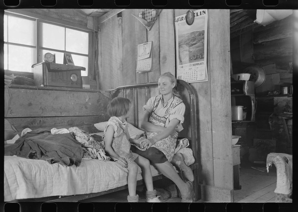 Black and white photo of a woman and young child sitting on a bed in a sparse rural cabin interior with wooden walls, calendars, a radio, and kitchen items visible in the background.